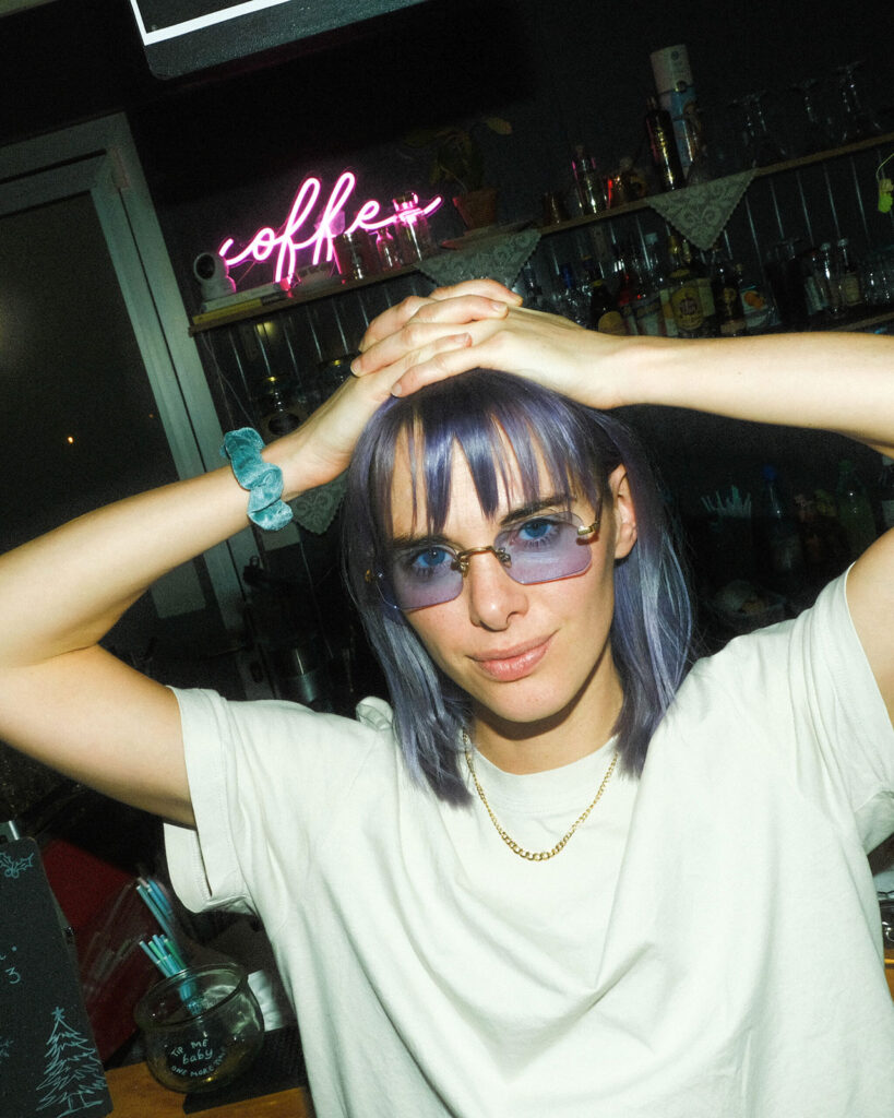 Frau mit getönter Designerbrille steht nachts vor einem Geschäft mit Neon-Schriftzug „coffee“